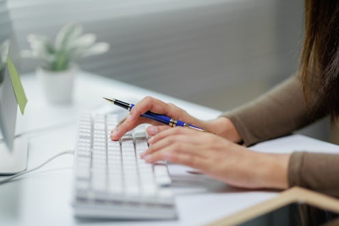 Close-up of female hands typing on a white keyboard while holding a blue pen, in a brightly lit offi...