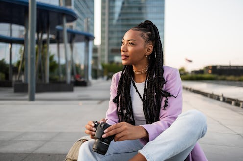 Young photographer sitting on the ground in a vibrant modern city, holding a camera while looking aw...