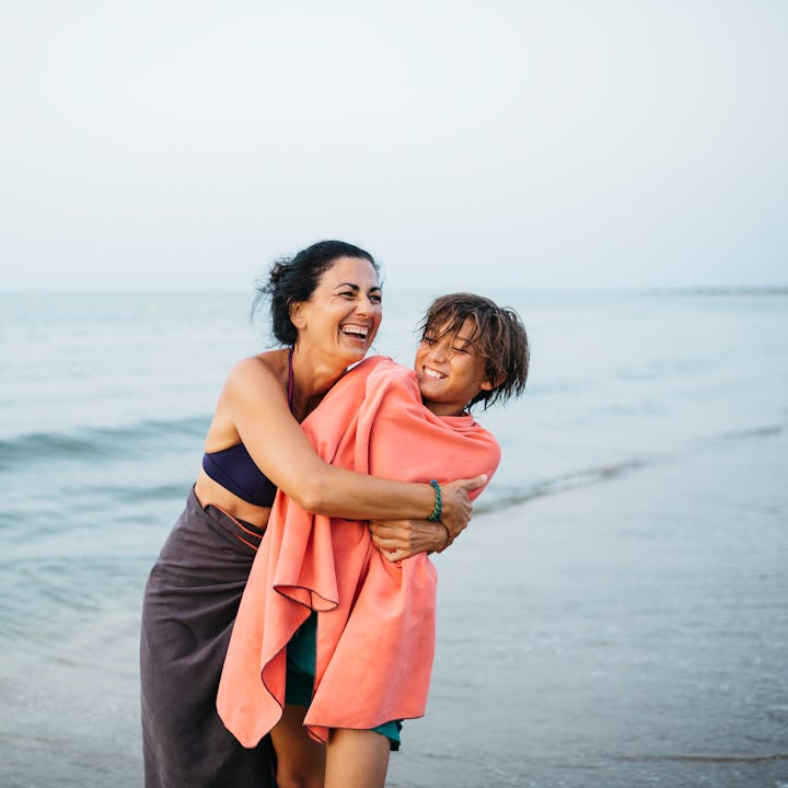 Portrait of a smiling mature woman and her twelve years old boy, embracing on the beach during sunse...