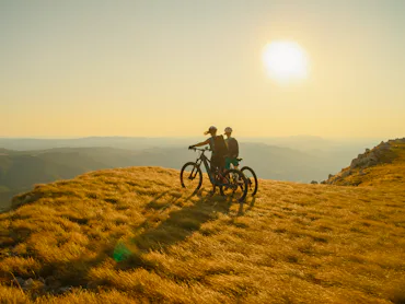 Two Female Cyclists Stand with their Mountain Bikes on a Grassy Mountaintop,Enjoying the Panoramic v...