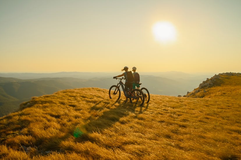 Two Female Cyclists Stand with their Mountain Bikes on a Grassy Mountaintop,Enjoying the Panoramic v...