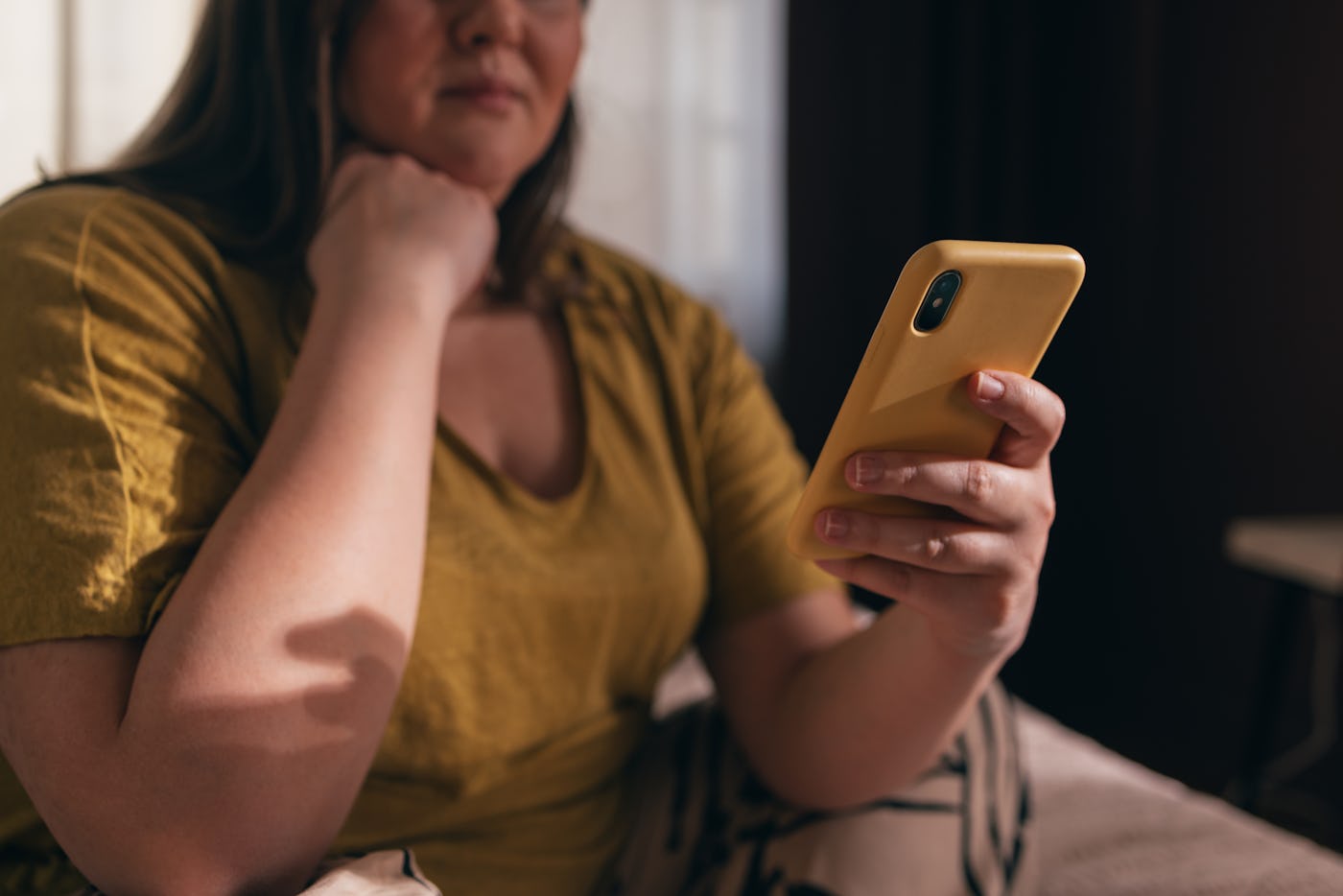 An unrecognizable plus size woman in a yellow T-shirt watching reels on her mobile phone.