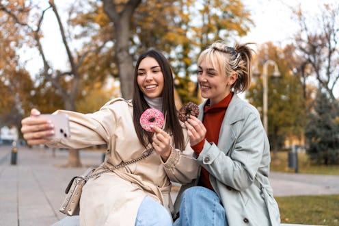 Two smiling young women enjoying donuts while taking a selfie with a smartphone in a vibrant park, s...