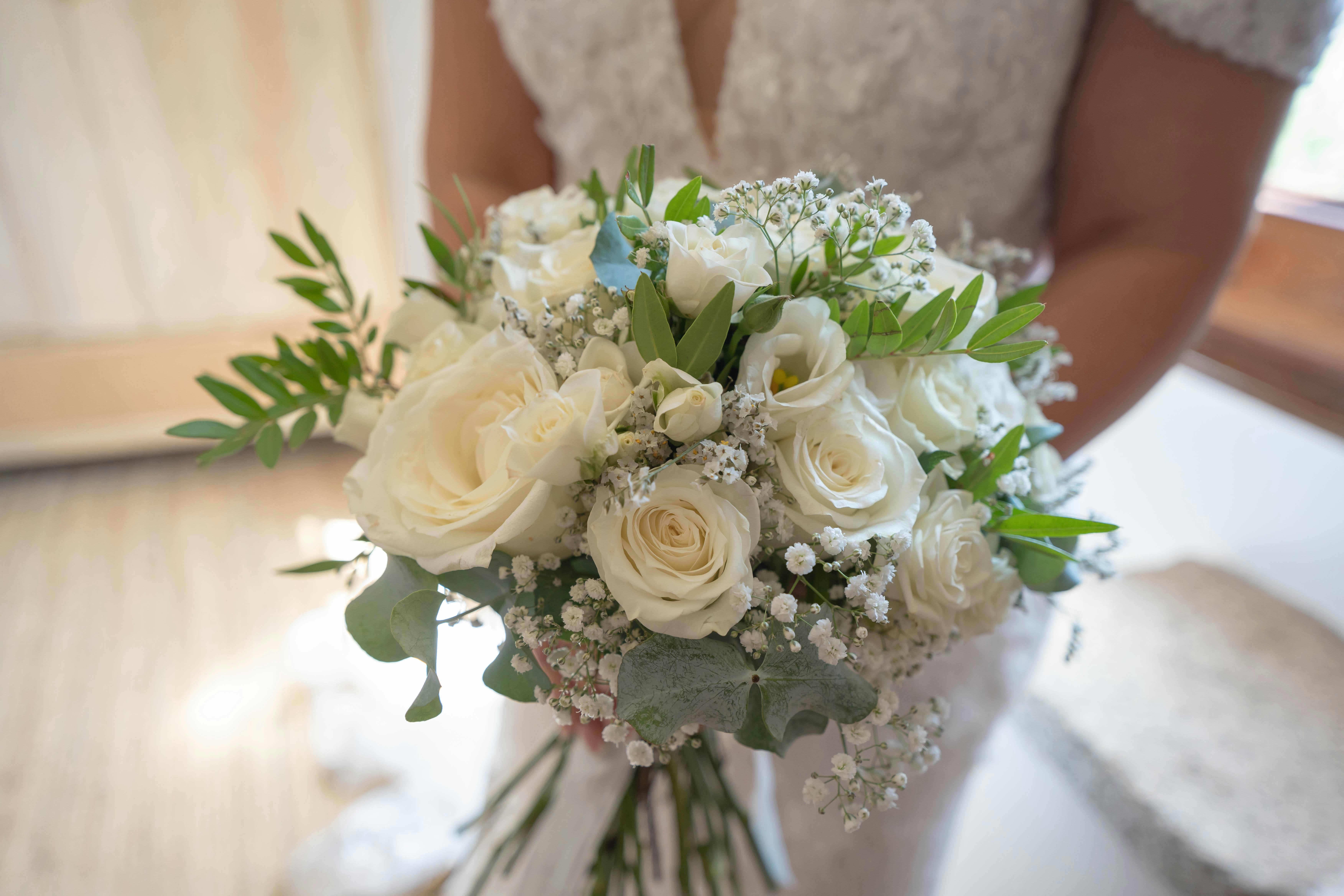 A close-up of a bride holding a beautifully arranged bouquet of white roses, baby's breath, and gree...