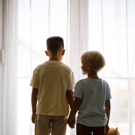 Two young brothers are holding hands and looking out the window, one of them holding a teddy bear