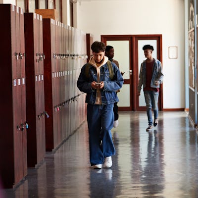 Teenage boy using smart phone while walking by lockers in front of friends at high school corridor