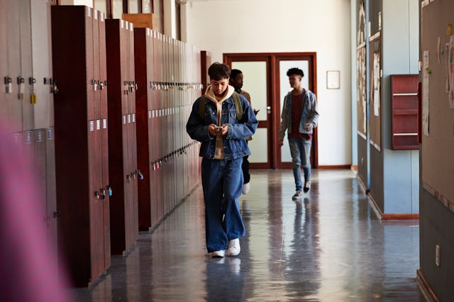Teenage boy using smart phone while walking by lockers in front of friends at high school corridor