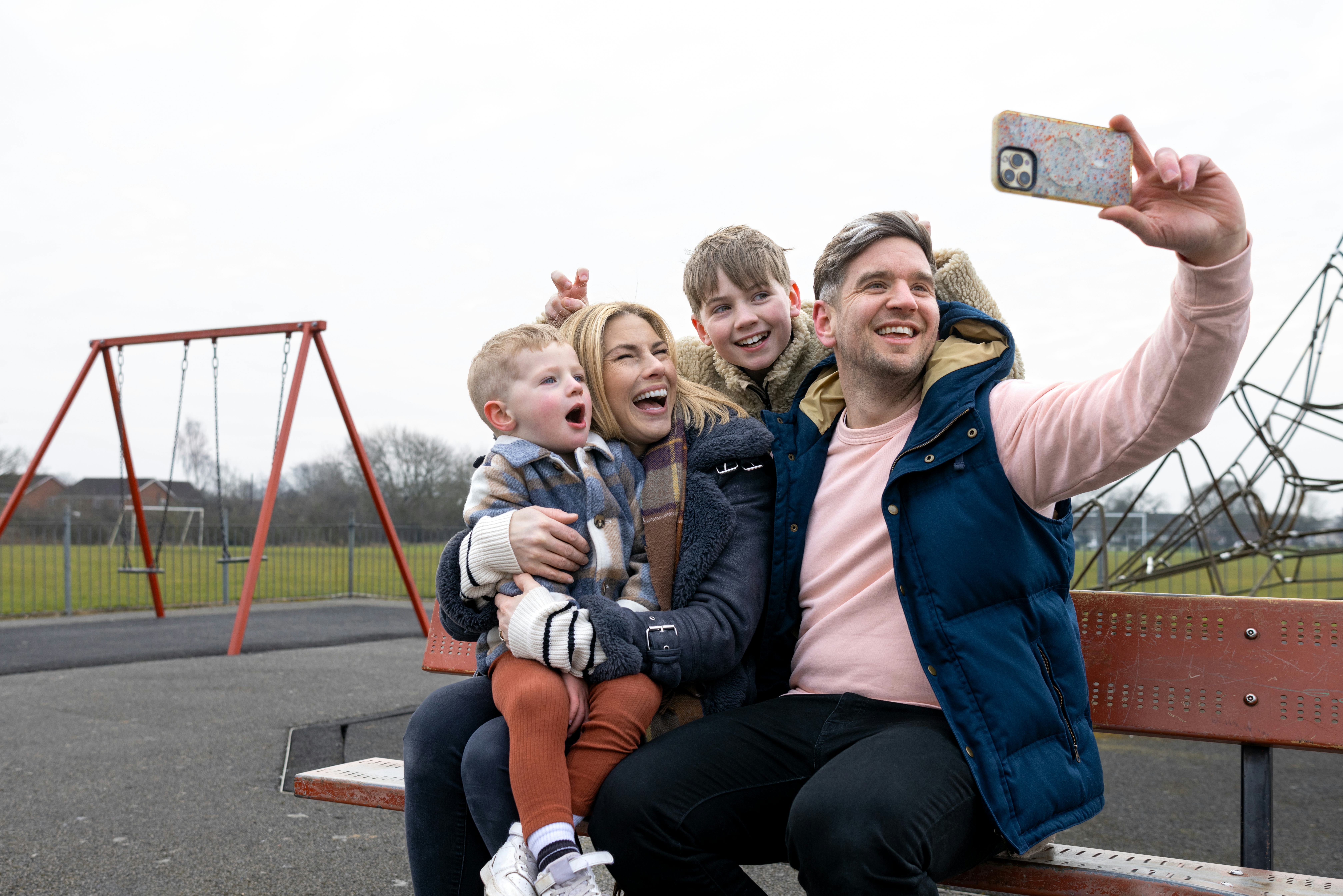 A wide shot of a mother and father sitting down on a bench with their children at a public park in D...