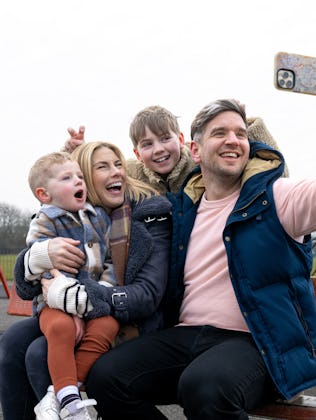 A wide shot of a mother and father sitting down on a bench with their children at a public park in D...
