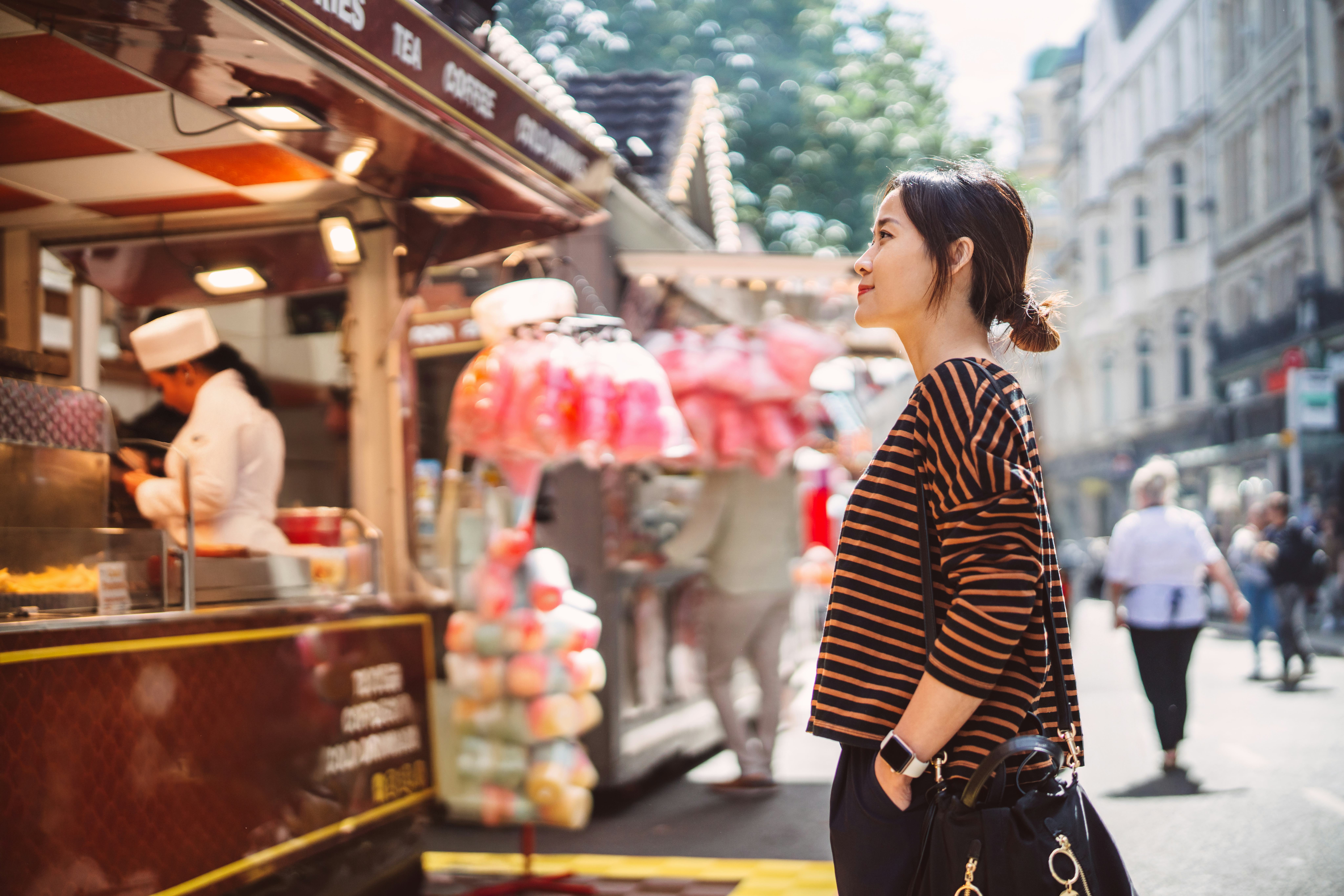 A woman pauses to look at a food stall while walking through a bustling street fair, surrounded by b...