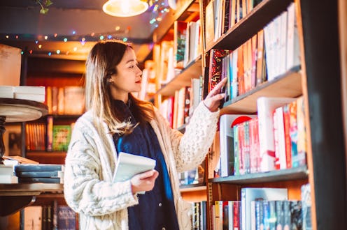 Asian woman browsing books in a warm, atmospheric bookstore, selecting a novel from a shelf while ho...