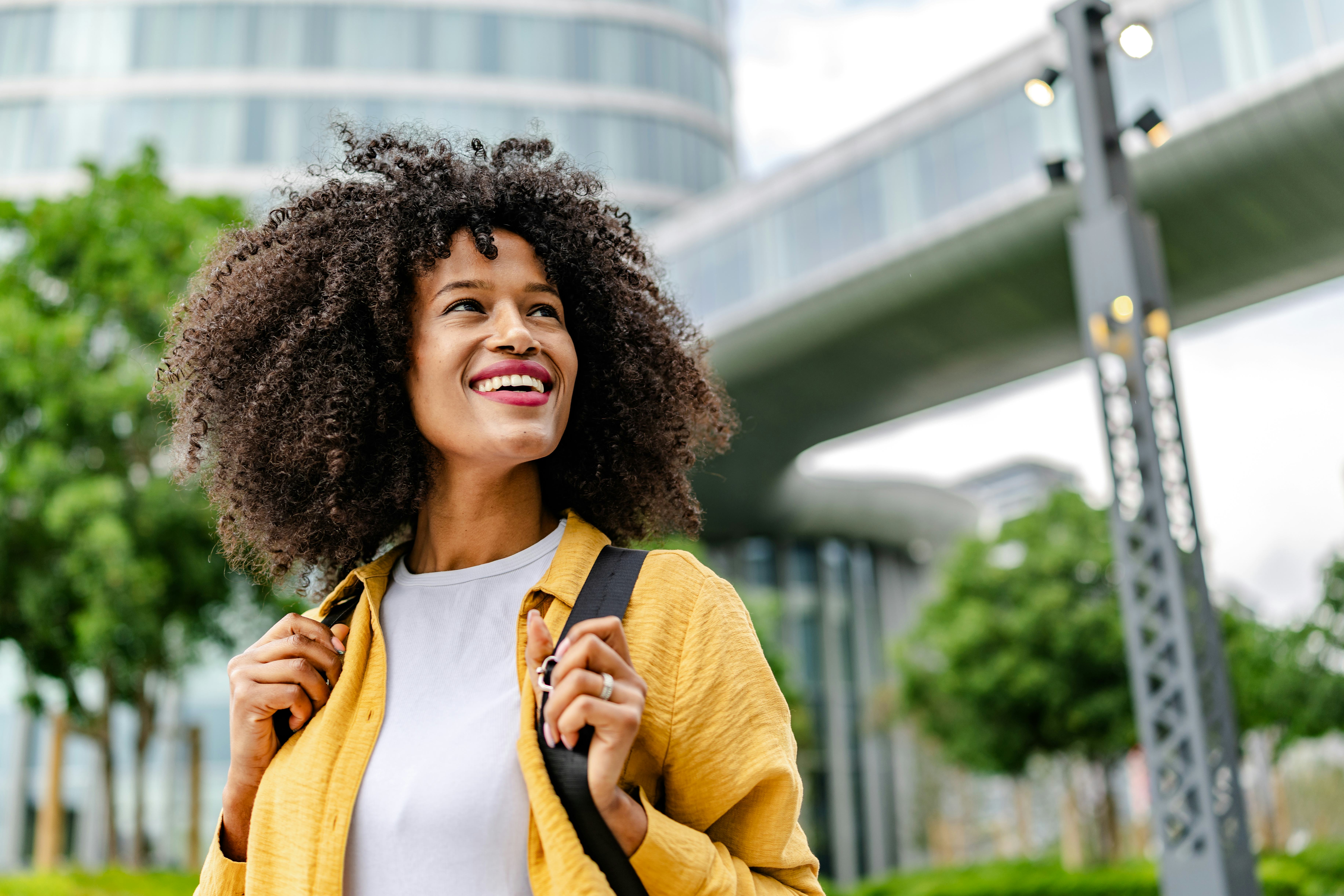 A young black woman stands outside and smiles brightly. She enjoys the day surrounded by tall buildi...