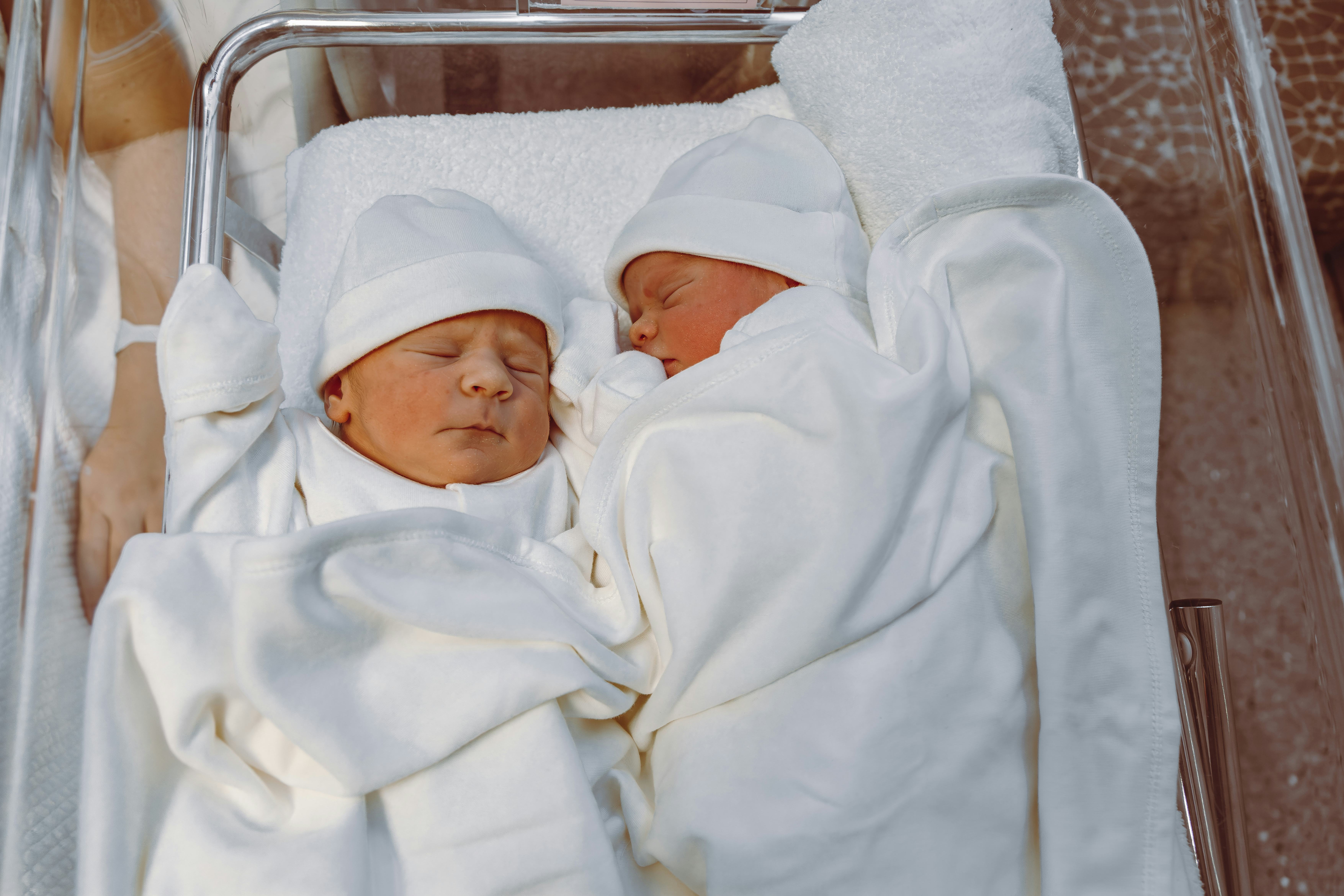 A serene image of newborn twins dressed in white swaddling clothes, lying side by side in a hospital...
