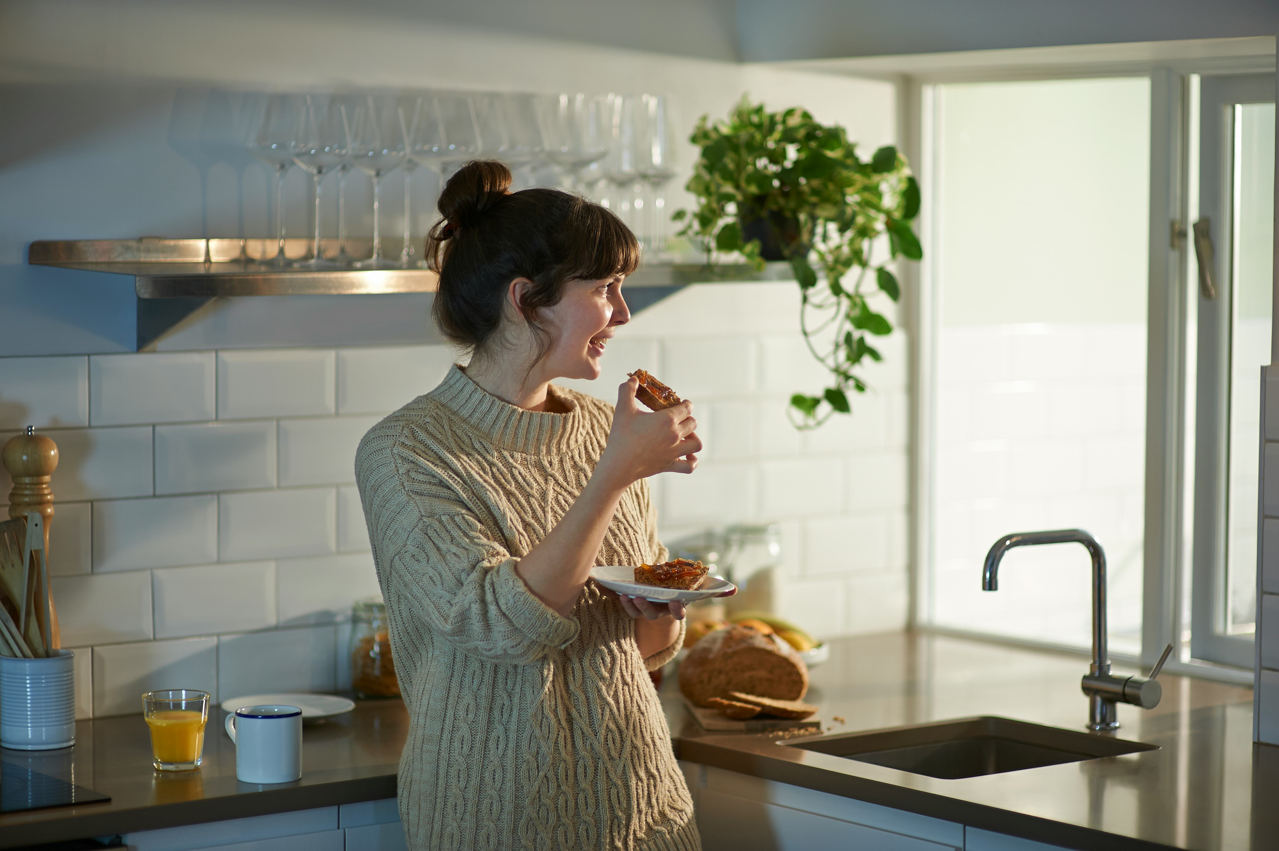 A happy and positive woman smiles as she eats her breakfast toast and looks out of the window in her...