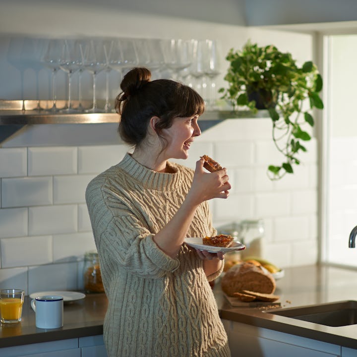 A happy and positive woman smiles as she eats her breakfast toast and looks out of the window in her...