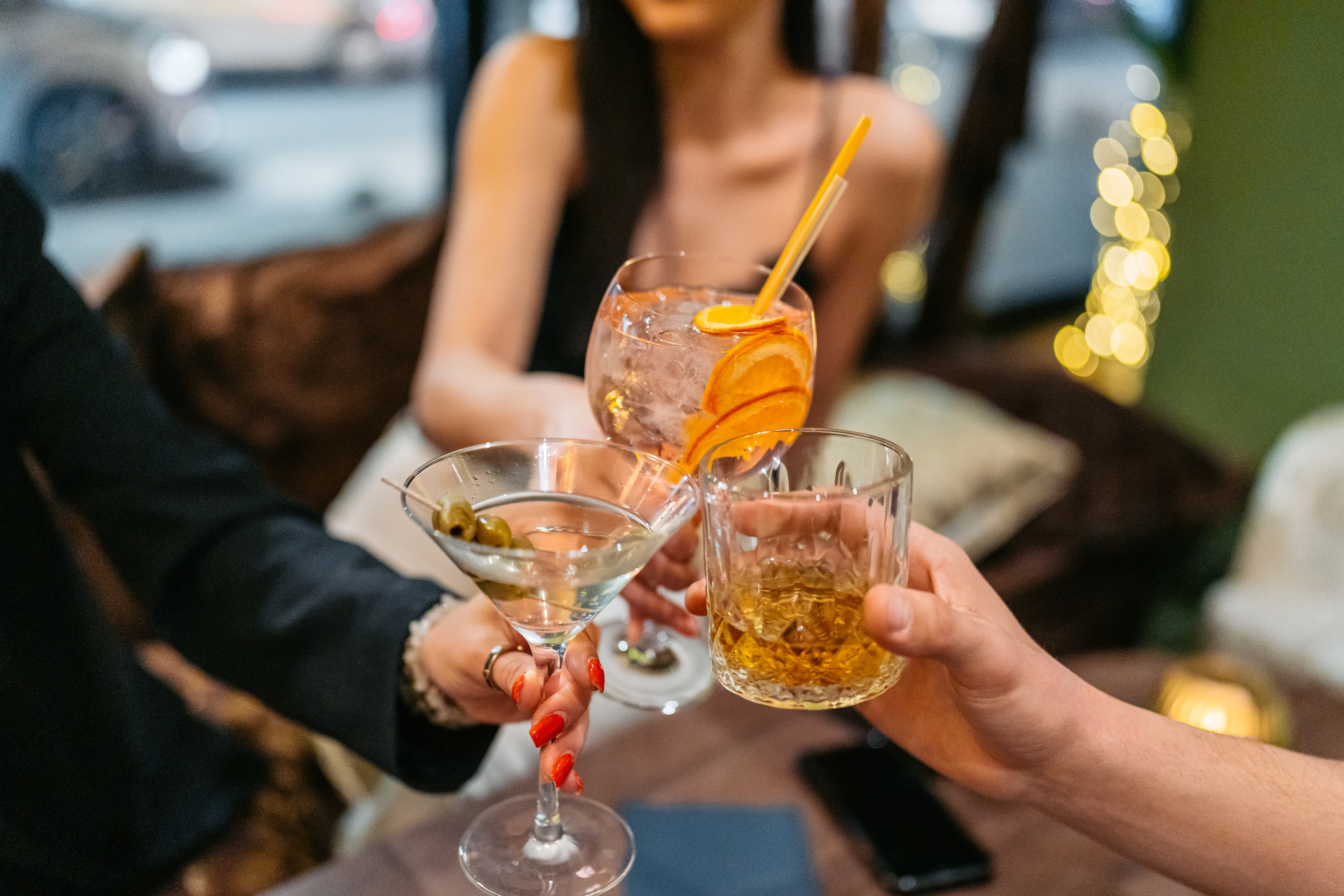 Group of three friends having a celebratory toast with cocktails and whiskey in a bar at night. Clos...