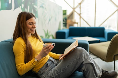 Young woman in a yellow sweater relaxes on a blue couch in a modern apartment, smiling as she sips c...