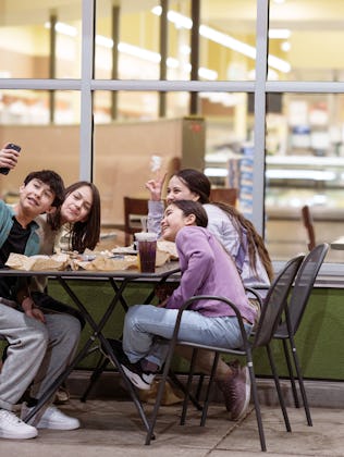 A cheerful group of Eurasian teens and tweens eat fast food in an outdoor dining area at a strip mal...