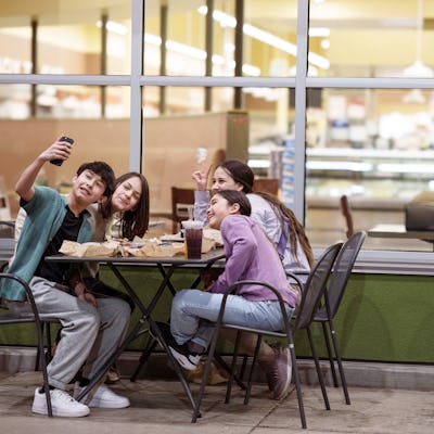 A cheerful group of Eurasian teens and tweens eat fast food in an outdoor dining area at a strip mall at night, and use a smart phone to take a selfie together.