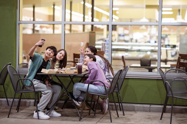 A cheerful group of Eurasian teens and tweens eat fast food in an outdoor dining area at a strip mal...