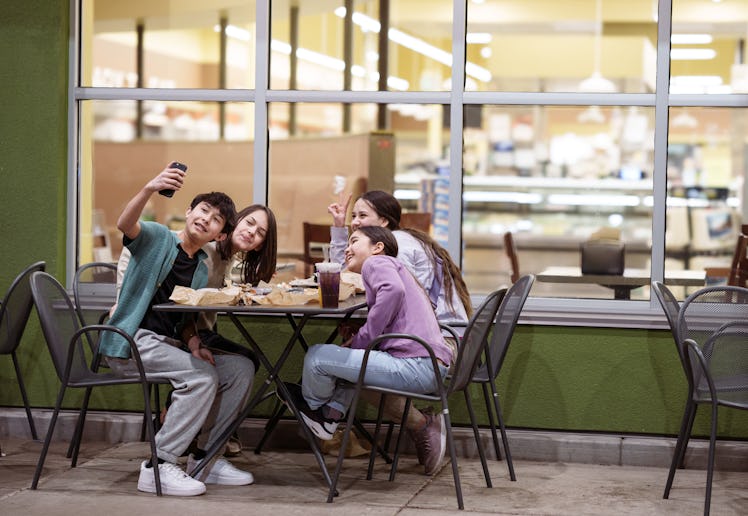 A cheerful group of Eurasian teens and tweens eat fast food in an outdoor dining area at a strip mal...