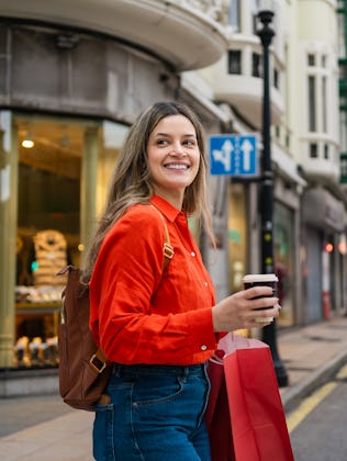 Young happy woman walking in city, holding shopping bags and disposable coffee cup. Looking joyful o...