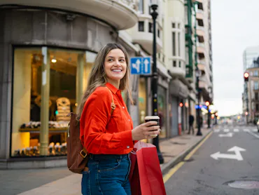 Young happy woman walking in city, holding shopping bags and disposable coffee cup. Looking joyful o...