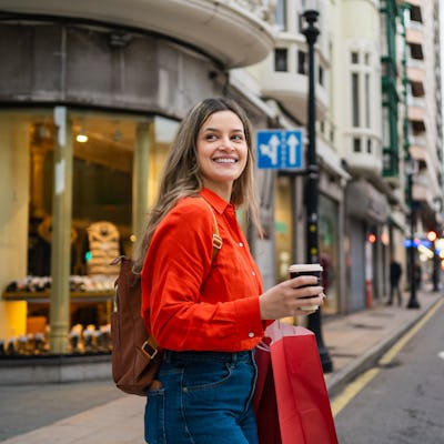 Young happy woman walking in city, holding shopping bags and disposable coffee cup. Looking joyful on urban street