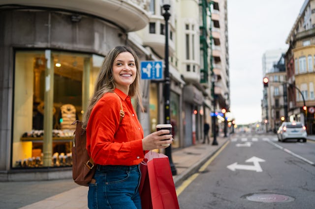 Young happy woman walking in city, holding shopping bags and disposable coffee cup. Looking joyful o...
