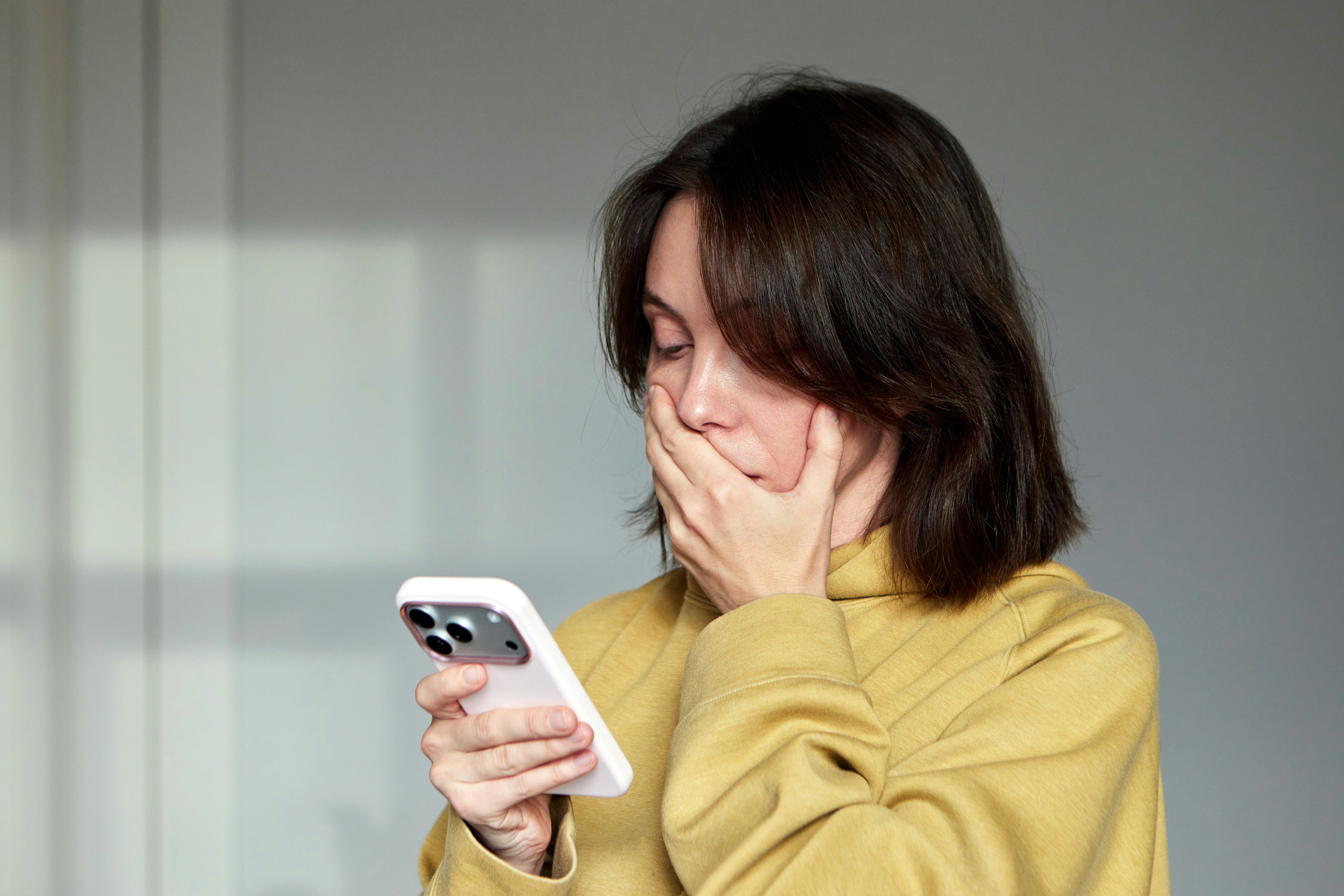 Woman holding a mobile phone and looking shocked or overwhelmed, covering her mouth with her hand af...