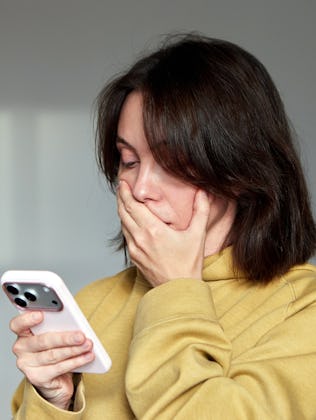 Woman holding a mobile phone and looking shocked or overwhelmed, covering her mouth with her hand af...