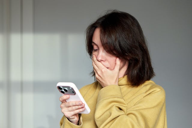 Woman holding a mobile phone and looking shocked or overwhelmed, covering her mouth with her hand af...