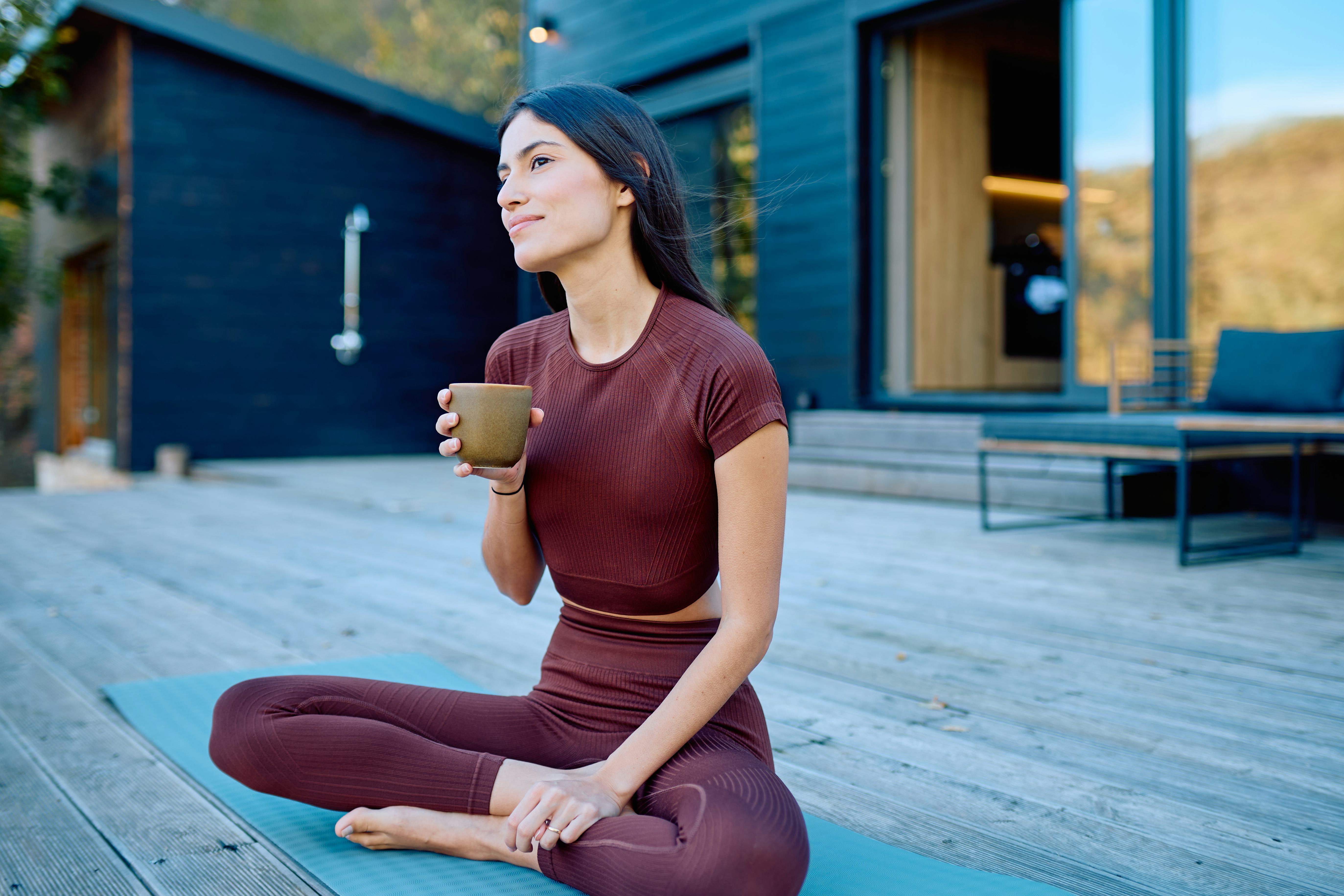 Young adult woman relaxing on a yoga mat, holding a coffee cup, and looking away thoughtfully in her...