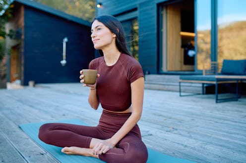 Young adult woman relaxing on a yoga mat, holding a coffee cup, and looking away thoughtfully in her...