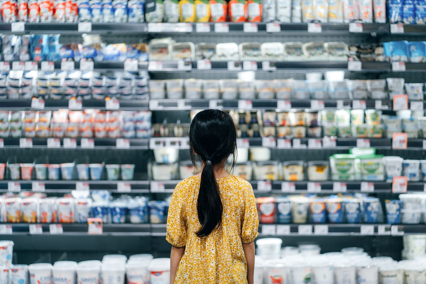 Rear view of Asian girl standing in front of a shelf full of fresh dairy products in the dairy aisle in a supermarket. Healthier food choice for kids. Healthy eating and well balanced diet. Concept of customer awareness
