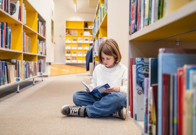 Young Caucasian boy sitting cross-legged on the library floor, deeply engrossed in a book. Surrounde...