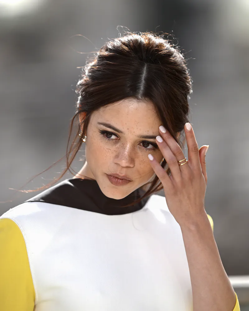Jenna Ortega moving her hair out of her face, her lips with a greigey nude color