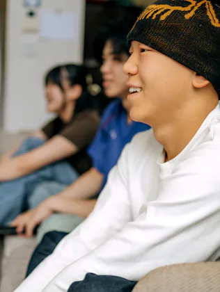 Three young friends sit on a sofa in a cozy living room, cheering and enjoying a sports game togethe...