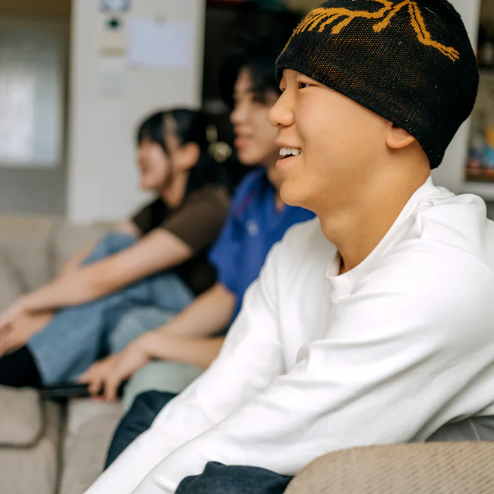 Three young friends sit on a sofa in a cozy living room, cheering and enjoying a sports game togethe...
