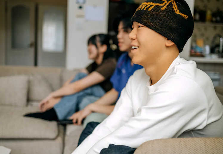 Three young friends sit on a sofa in a cozy living room, cheering and enjoying a sports game togethe...