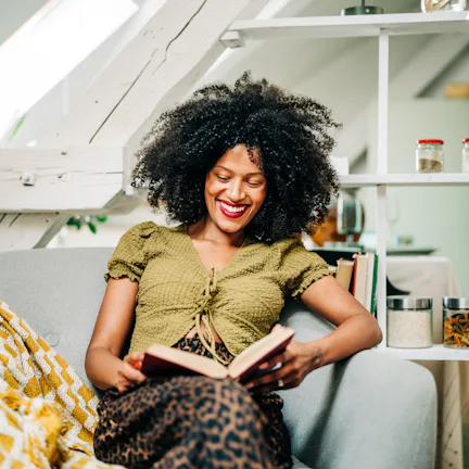 A young woman drinks tea while relaxing and reading a book in the living room.