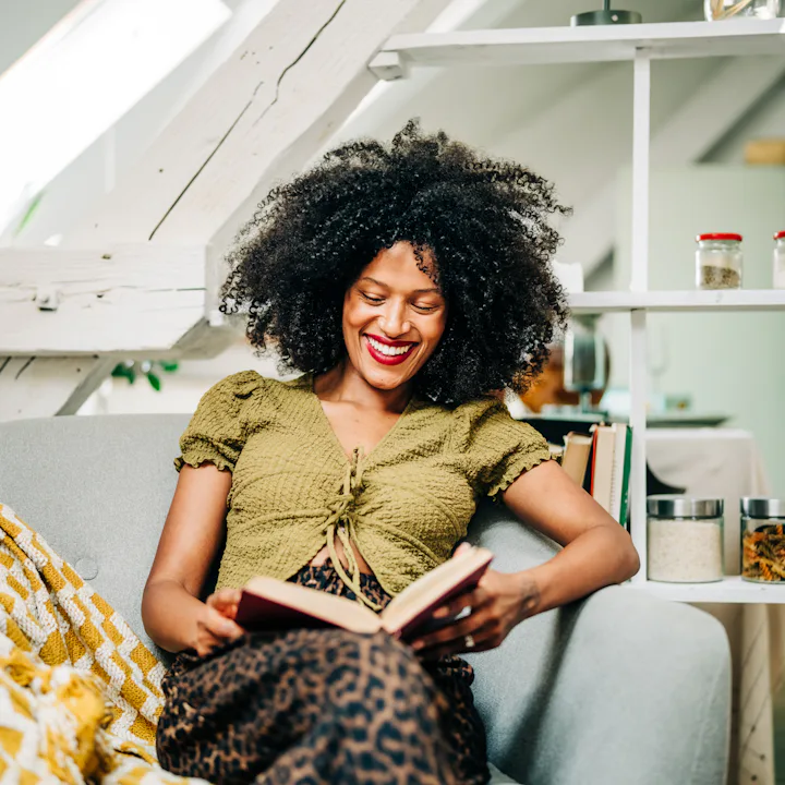 A young woman drinks tea while relaxing and reading a book in the living room.