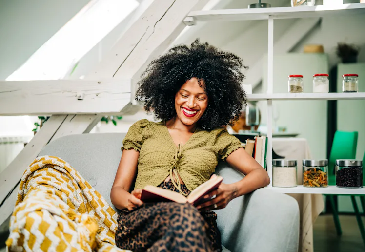 A young woman drinks tea while relaxing and reading a book in the living room.