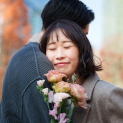 Young woman gently holds a bouquet of flowers while leaning into her partner, smiling with her eyes ...