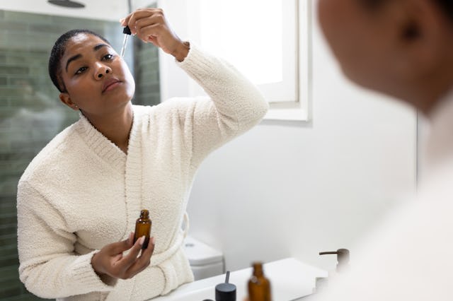 Young woman wearing bathrobe using cosmetic serum in front of bathroom mirror during her morning rou...