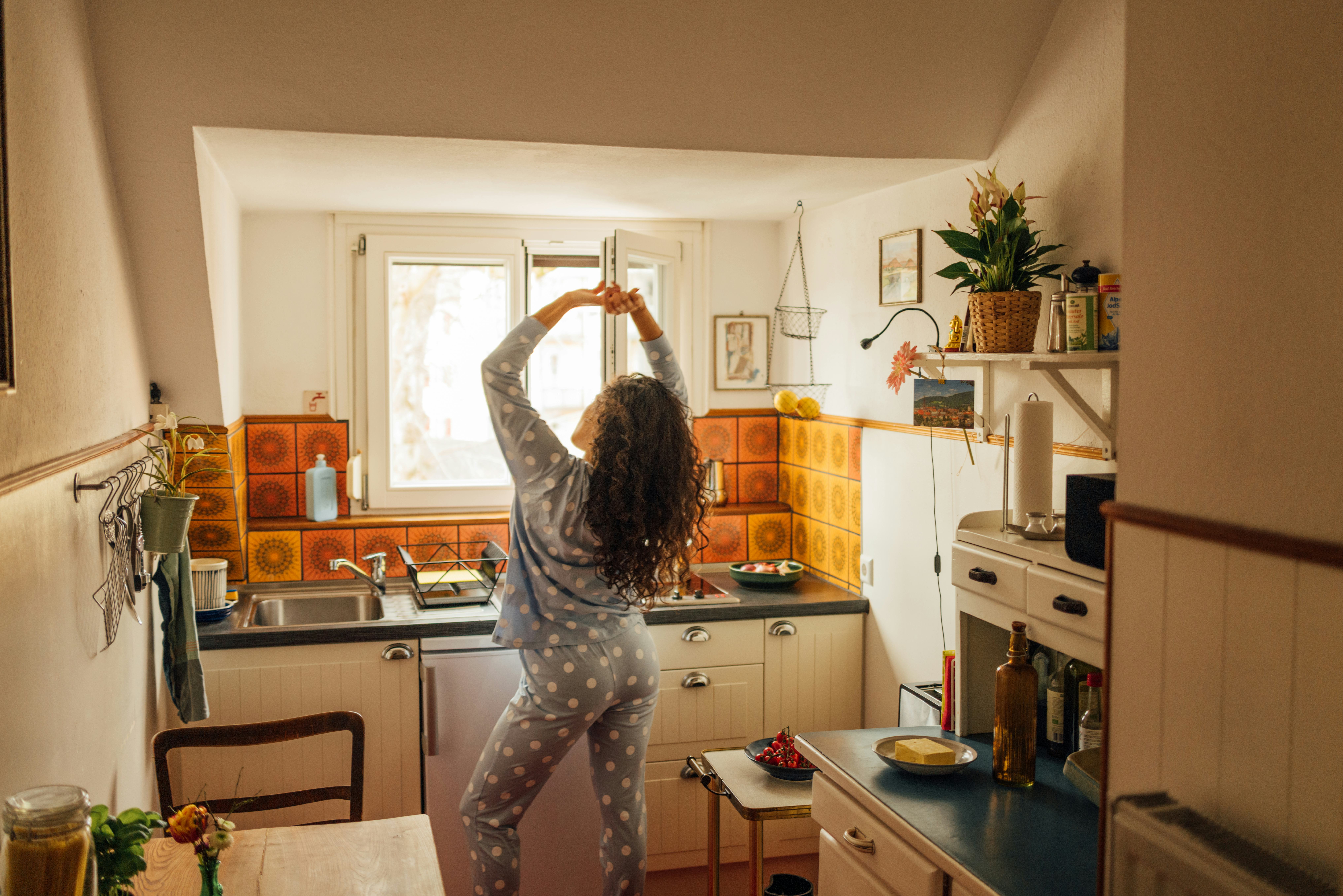 A woman in polka dot pajamas stretches by the kitchen window, enjoying a quiet morning. Warm light a...