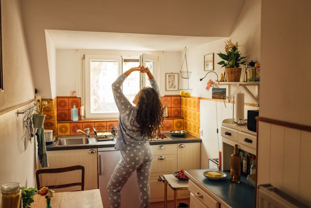 A woman in polka dot pajamas stretches by the kitchen window, enjoying a quiet morning. Warm light a...