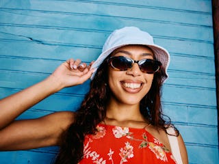 Closeup shot of a smiling young woman in her 20s wearing sunglasses and a light summer hat, standing...