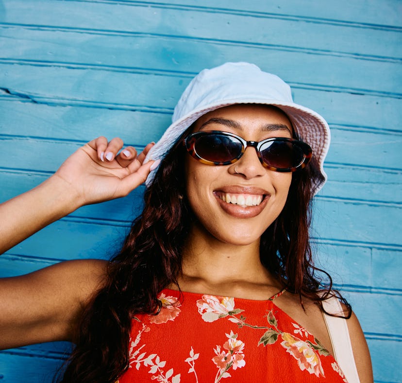 Closeup shot of a smiling young woman in her 20s wearing sunglasses and a light summer hat, standing...