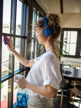 Photo of young female cleaning window in her apartment with headphones.